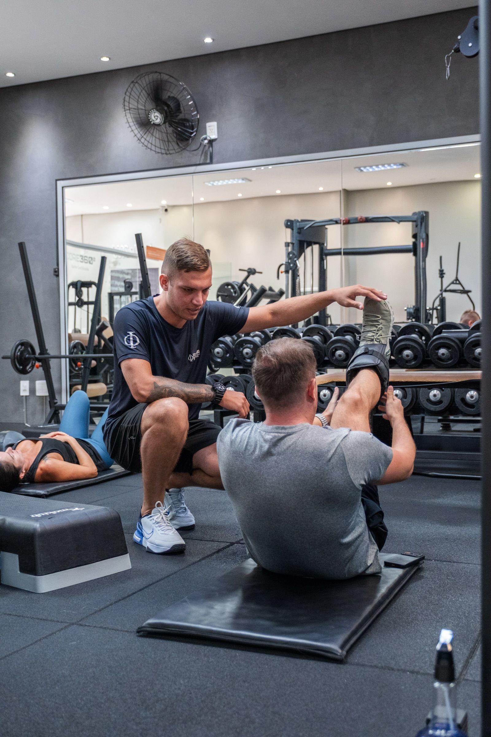 Professor corrigindo um aluno no exercício de abdominal, com os halteres e os aparelhos do studio ao fundo